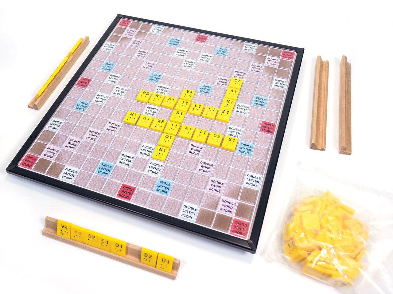 Tablero de Scrabble en versión braille con fichas amarillas que combinan la letra impresa, su valor numérico y los puntos braille en relieve; a los lados, atriles de madera con fichas y bolsa de repuesto.